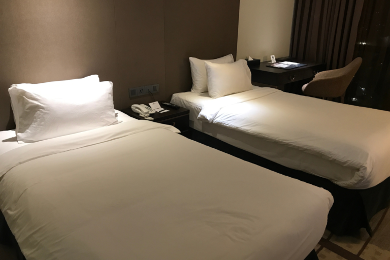 Comfortable hotel room with twin beds, clean white linens, bedside table, and work desk setup
