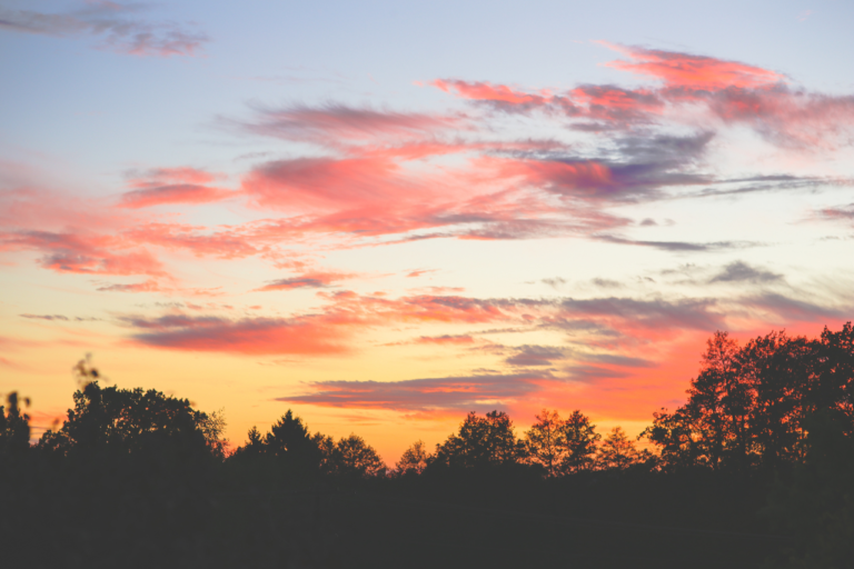 Colorful sunset sky with pink clouds over silhouetted trees, symbolizing peaceful travel moments and reflection during a journey.
